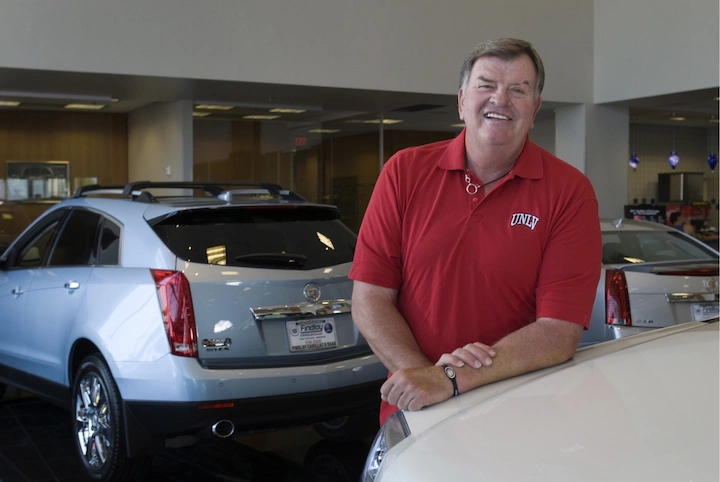 Cliff Findlay smiling inside his car dealership, standing near luxury vehicles, wearing red UNLV shirt.