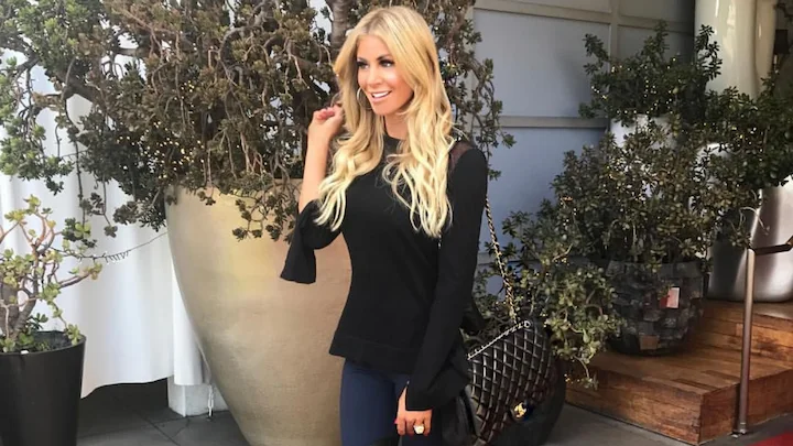 Jackie Melby smiling in black outfit with handbag, standing outdoors near plants and large pots.