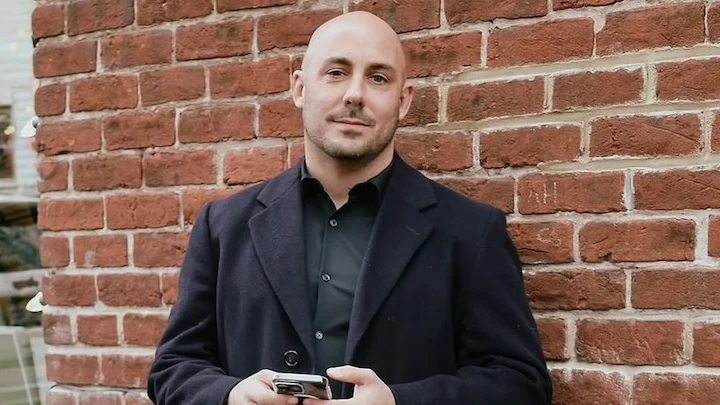Man in dark coat holding a smartphone, standing against a red brick wall, urban background.